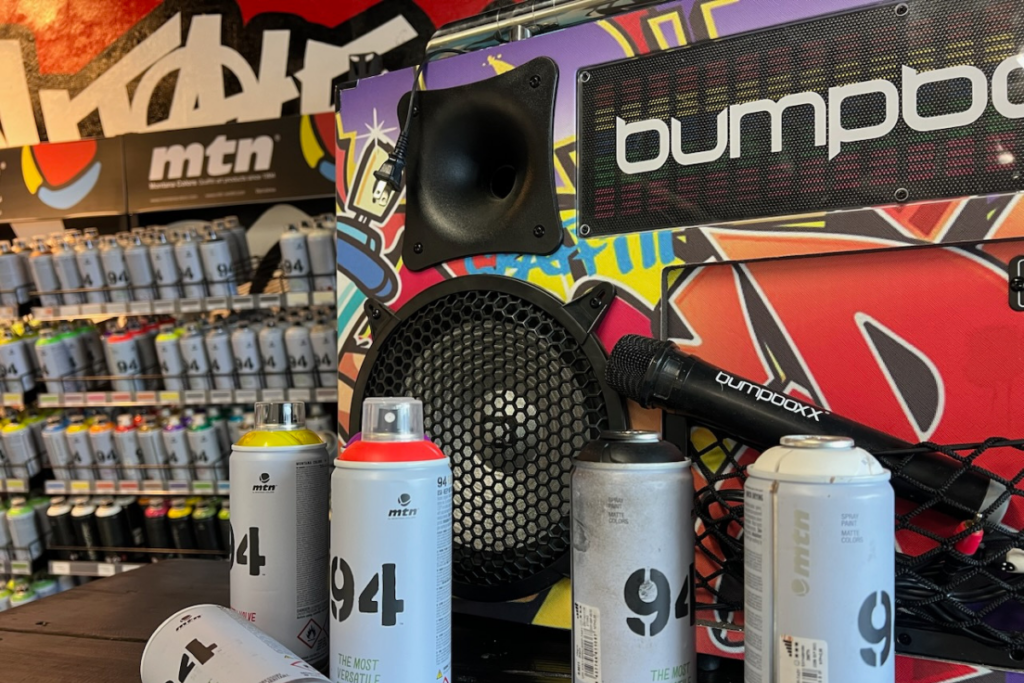 Colorful graffiti art setup featuring spray paint cans and a Bumpboxx speaker at Overspray Art Space.